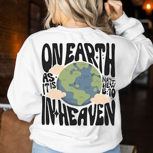 May include: White sweatshirt with a black graphic design featuring a stylised earth with clouds and the text "On Earth As It Is In Heaven" with the scripture reference "Matthew 6:10".