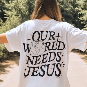 以下が含まれることがあります： 白いTシャツに、黒いグラフィックで「OUR WORLD NEEDS JESUS」と、スタイリッシュな地球儀と星が描かれています。
