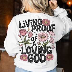 Puede incluir: Sudadera blanca con el texto "Living Proof of a Loving God" en letras negras con flores rosas y hojas verdes.