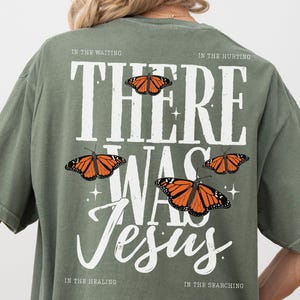 May include: An olive green t-shirt featuring a large white graphic on the back. The text reads "THERE WAS JESUS" with four monarch butterflies and the phrases "IN THE WAITING," "IN THE HURTING," "IN THE HEALING," and "IN THE SEARCHING" printed.