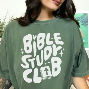May include: A green t-shirt with a white graphic that says "Bible Study Club" in a retro font. The graphic also includes a small heart and a stylized image of a closed book with a cross on the cover.