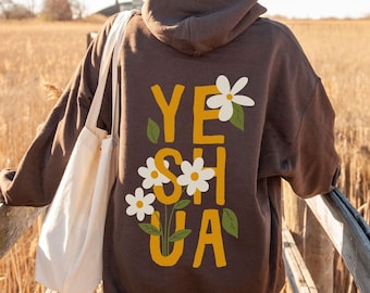 以下が含まれることがあります： 黄色のグラフィックで「YESHUA」と書かれた茶色のフード付きスウェットシャツ。白い花と緑の葉が描かれています。
