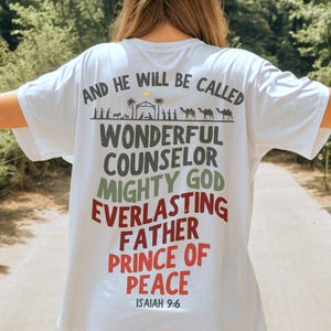 May include: A white short-sleeve t-shirt featuring a graphic on the back with the text AND HE WILL BE CALLED WONDERFUL COUNSELOR MIGHTY GOD EVERLASTING FATHER PRINCE OF PEACE ISAIAH 9:6 and a silhouette illustration of a nativity scene with camels.