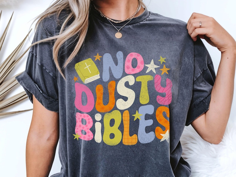 May include: Dark gray t-shirt with the text "NO DUSTY BIBLES" in colorful, playful lettering. The design includes a book and stars. The shirt has a vintage, worn look.