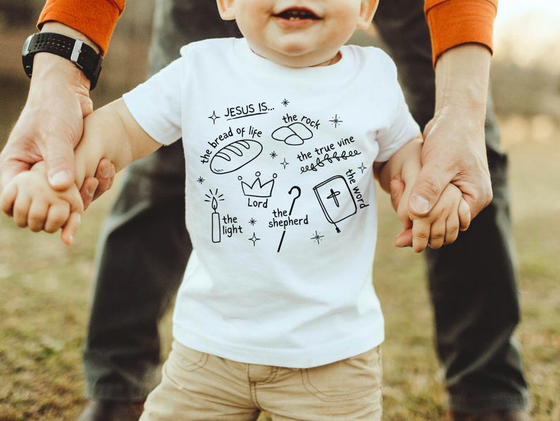 May include: White toddler t-shirt with black ink illustrations of a candle, bread, crown, vine, rock, book, and a question mark. The text on the shirt reads "JESUS IS... the bread of life the light Lord the rock the true vine the word the shepherd".