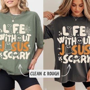 May include: An olive green and a charcoal grey t-shirt, each featuring the cream-colored text "LIFE WITHOUT JESUS IS SCARY" with orange stitching details on the word "JESUS". The design includes a skull, spiderweb, ghost, and bat graphic.