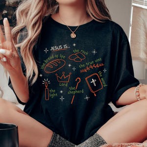 May include: A black t-shirt with a white graphic design that says "JESUS IS..." and lists several attributes of Jesus, including "the bread of life", "the rock", "the true vine", "the light", "the Lord", "the shepherd", and "the word".