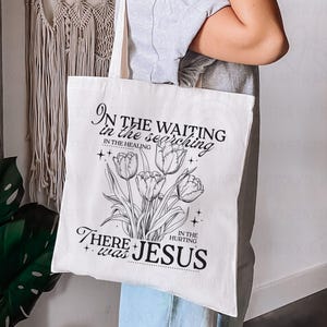 May include: Cream-colored tote bag featuring a black and white floral design with the text "In the waiting, in the searching, in the healing, in the hurting, there was Jesus."