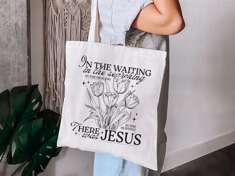 May include: Cream-colored tote bag featuring a black and white floral design with the text "In the waiting, in the searching, in the healing, in the hurting, there was Jesus."