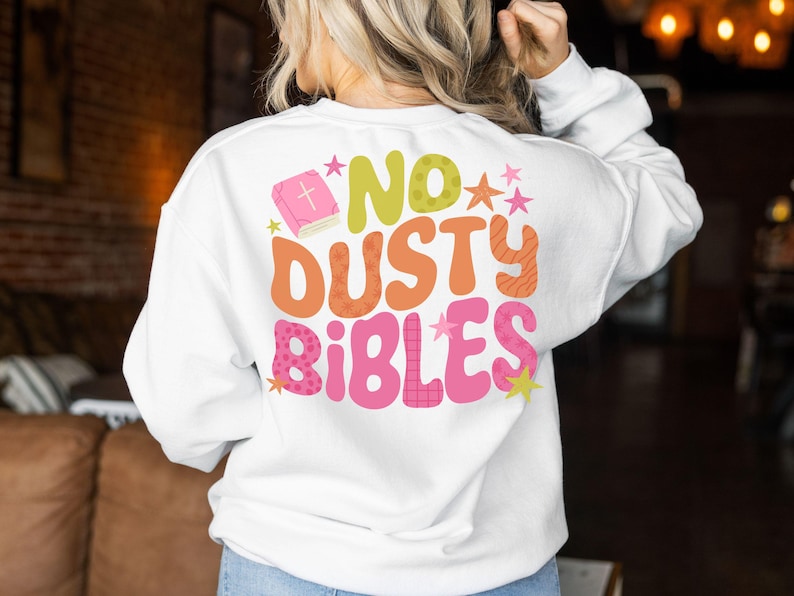 May include: White sweatshirt with the text "NO DUSTY BIBLES" in a retro font. The design includes a pink book, stars, and a mix of orange, green, and pink colors. A casual, comfortable top.