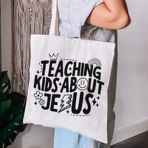 Puede incluir: Un bolso de mano blanco con el texto "TEACHING KIDS ABOUT JESUS" en negro, con una corona, un arco&iacute;ris, una carita sonriente, una flor y gr&aacute;ficos de rel&aacute;mpagos. El bolso lo sostiene una persona.
