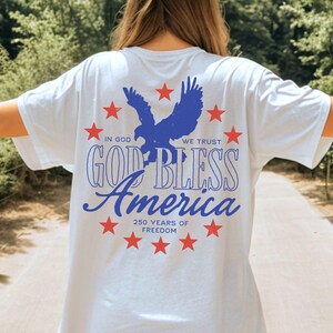 May include: A white t-shirt featuring a blue eagle graphic surrounded by red stars. The text reads IN GOD WE TRUST, GOD BLESS AMERICA, and 250 YEARS OF FREEDOM. The design is printed on the back of the shirt, showcasing patriotic colors and themes.