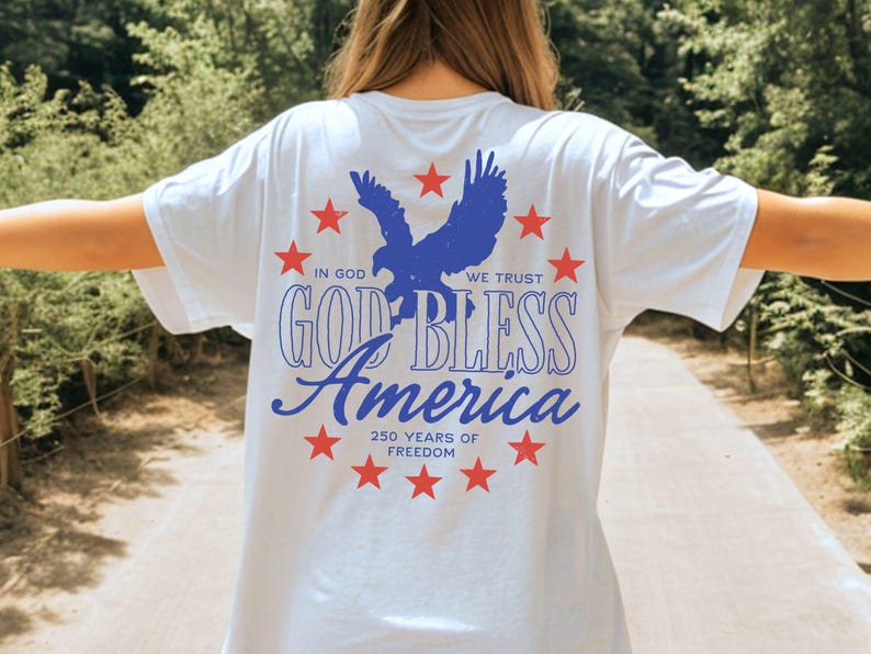 May include: A white t-shirt featuring a blue eagle graphic surrounded by red stars. The text reads IN GOD WE TRUST, GOD BLESS AMERICA, and 250 YEARS OF FREEDOM. The design is printed on the back of the shirt, showcasing patriotic colors and themes.