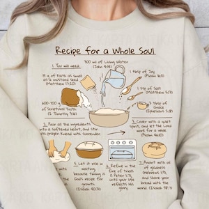Puede incluir: Sudadera beige con un gráfico que ilustra una receta para un alma completa, incluyendo ingredientes como fe, verdad y gracia. La receta está escrita en texto negro con ilustraciones de los ingredientes y pasos.