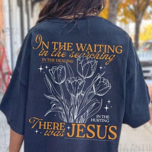 May include: Navy blue t-shirt with a floral design and the text "In the waiting, in the searching, in the healing, in the hurting, there was Jesus." The design features a bouquet of tulips. The shirt is a comfortable, casual style.