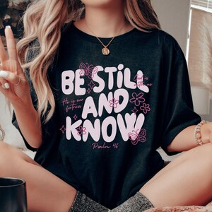 May include: Black t-shirt with the words "BE STILL AND KNOW" in a light pink, stylized font. The shirt also features floral and butterfly designs, along with the text "He is our fortress" and "Psalm 46".