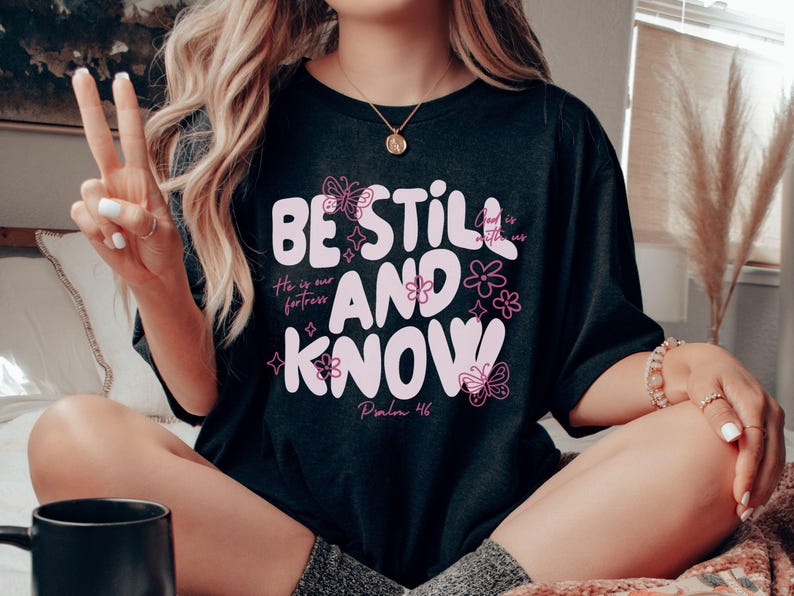 May include: Black t-shirt with the words "BE STILL AND KNOW" in a light pink, stylized font. The shirt also features floral and butterfly designs, along with the text "He is our fortress" and "Psalm 46".