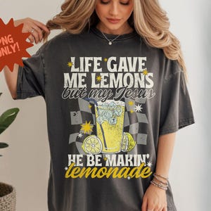 May include: Dark gray t-shirt with the text "LIFE GAVE ME LEMONS but my Jesus HE BE MAKIN' lemonade." The graphic includes a glass of lemonade, lemons, and star accents. The shirt is a vintage style.