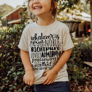 May include: A cream-colored t-shirt with black text that reads "whatever is true, noble, right, pure, lovely, admirable, praiseworthy, excellent, think about such things." The shirt is worn by a child.