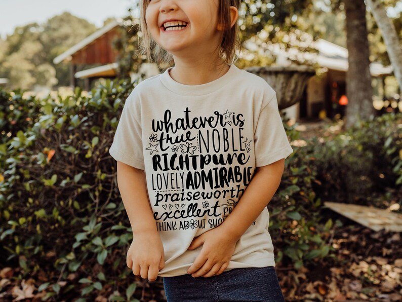 May include: A cream-colored t-shirt with black text that reads "whatever is true, noble, right, pure, lovely, admirable, praiseworthy, excellent, think about such things." The shirt is worn by a child.