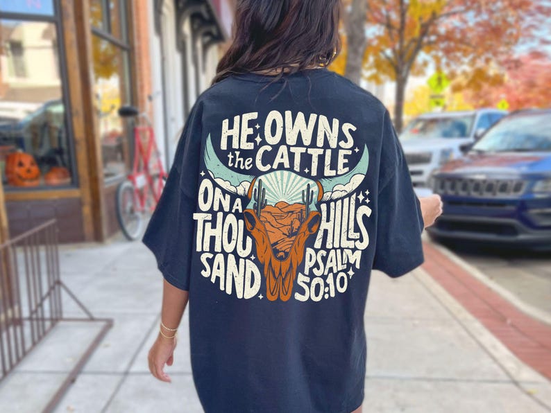 May include: Navy blue t-shirt with a graphic design on the back. The design features a bull skull, cacti, and the text "HE OWNS THE CATTLE ON A THOUSAND HILLS PSALM 50:10". The shirt is a casual wear item.
