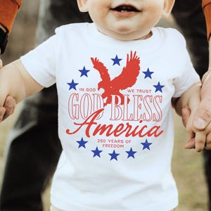 May include: A white short-sleeved t-shirt featuring a red eagle silhouette surrounded by a circle of blue stars. The text reads IN GOD WE TRUST, GOD BLESS America, and 250 YEARS OF FREEDOM in red and blue lettering on the front of the garment.