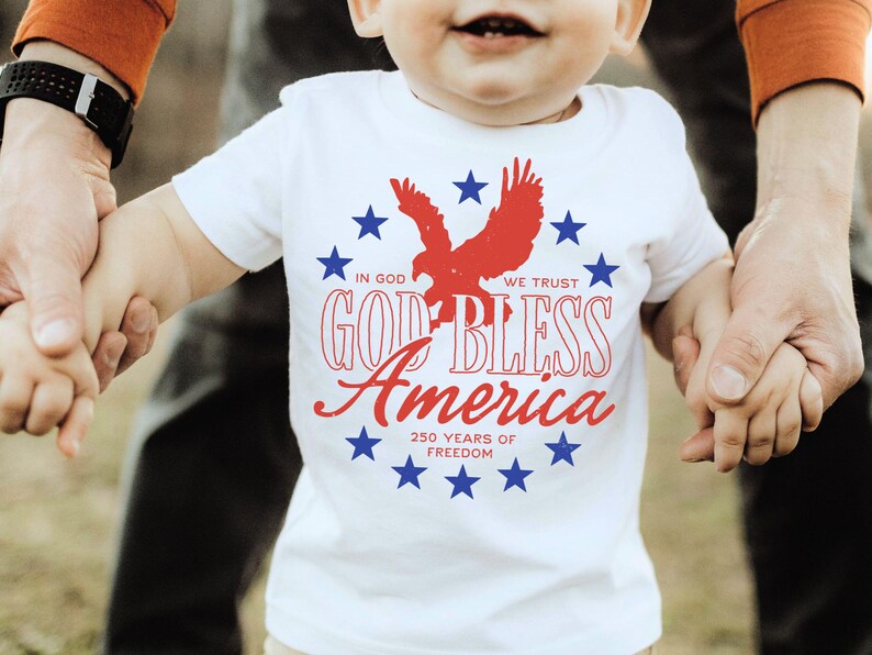 May include: A white short-sleeved t-shirt featuring a red eagle silhouette surrounded by a circle of blue stars. The text reads IN GOD WE TRUST, GOD BLESS America, and 250 YEARS OF FREEDOM in red and blue lettering on the front of the garment.