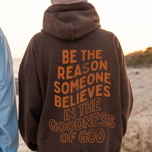 Może przedstawiać: Brązowa bluza z kapturem z nadrukiem "BE THE REASON SOMEONE BELIEVES IN THE GOODNESS OF GOD" w kolorze pomarańczowym na plecach.