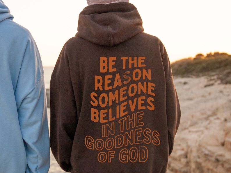 Może przedstawiać: Brązowa bluza z kapturem z nadrukiem "BE THE REASON SOMEONE BELIEVES IN THE GOODNESS OF GOD" w kolorze pomarańczowym na plecach.