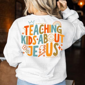 Puede incluir: Sudadera blanca con el texto "TEACHING KIDS ABOUT JESUS" en letras coloridas. El dise&ntilde;o incluye una corona, un arco&iacute;ris, una carita sonriente, una flor, un rayo y estrellas. La sudadera es de cuello redondo y manga larga.
