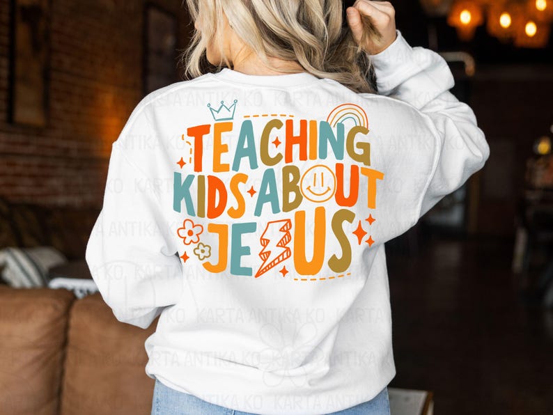 Puede incluir: Sudadera blanca con el texto "TEACHING KIDS ABOUT JESUS" en letras coloridas. El dise&ntilde;o incluye una corona, un arco&iacute;ris, una carita sonriente, una flor, un rayo y estrellas. La sudadera es de cuello redondo y manga larga.