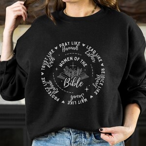 May include: Black sweatshirt with a white graphic design featuring a circle with the words "Women of the Bible" and a Bible with flowers. The circle is surrounded by phrases like "Trust like Ruth", "Pray like Hannah", "Lead like Esther", "Rejoice like Mary", "Wait like Sarah", "Protect like Ruth", and "Serve like Mary".