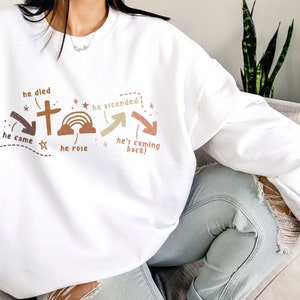 May include: White sweatshirt with a brown and green graphic design featuring a cross, rainbow, and arrows. The text on the graphic reads "he died", "he ascended", "he came", "he rose", and "he's coming back!"