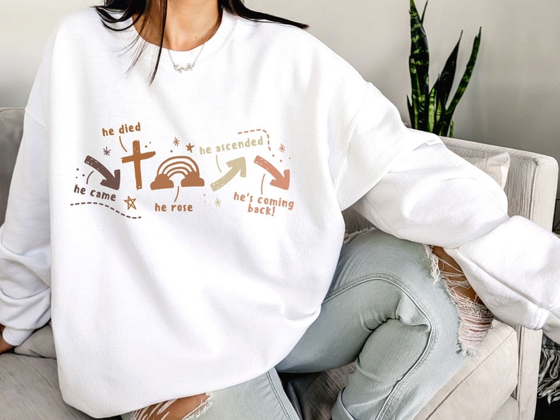May include: White sweatshirt with a brown and green graphic design featuring a cross, rainbow, and arrows. The text on the graphic reads "he died", "he ascended", "he came", "he rose", and "he's coming back!"