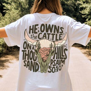 May include: White t-shirt with a desert-themed graphic on the back. The design features a cow skull, cacti, and text that reads "He owns the cattle on a thousand hills Psalm 50:10". The graphic is in shades of brown, pink, and green.