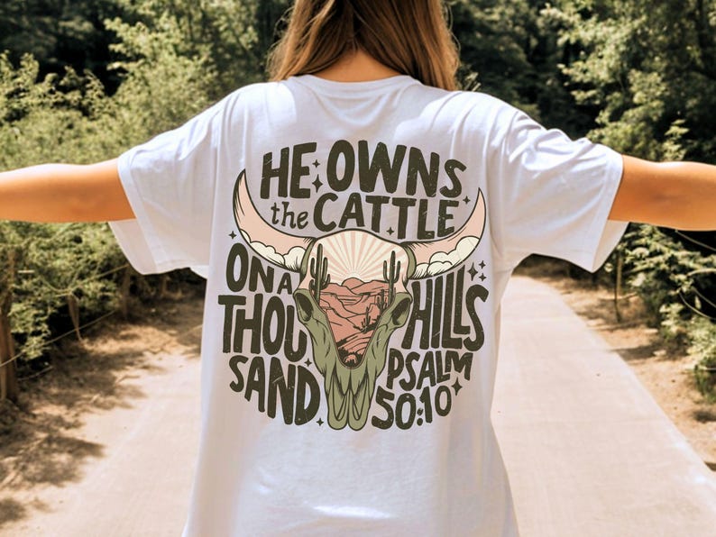 May include: White t-shirt with a desert-themed graphic on the back. The design features a cow skull, cacti, and text that reads "He owns the cattle on a thousand hills Psalm 50:10". The graphic is in shades of brown, pink, and green.