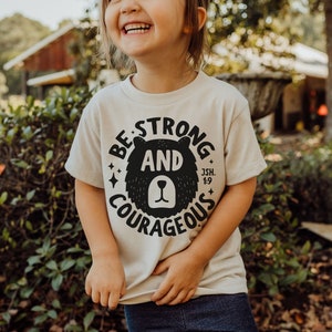 May include: A child wearing a beige t-shirt with a black bear graphic and the text "Be Strong and Courageous Jsh. 1:9".