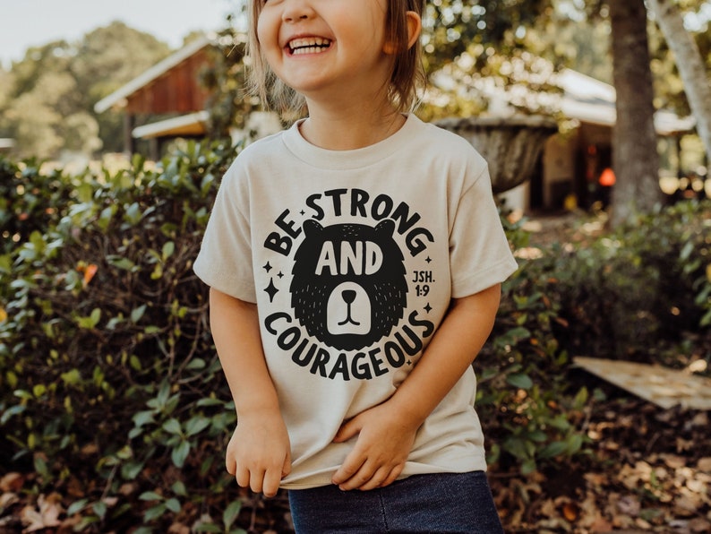 May include: A child wearing a beige t-shirt with a black bear graphic and the text "Be Strong and Courageous Jsh. 1:9".
