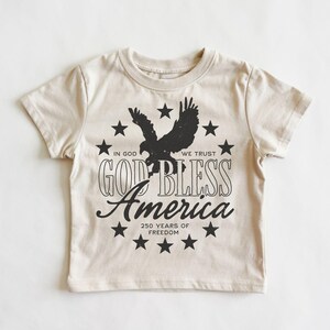 May include: A cream-colored short-sleeve t-shirt featuring a black graphic of a soaring eagle surrounded by a circle of stars. The text reads: IN GOD WE TRUST, GOD BLESS America, and 250 YEARS OF FREEDOM in a mix of block and script fonts.