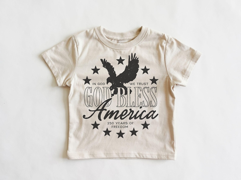May include: A cream-colored short-sleeve t-shirt featuring a black graphic of a soaring eagle surrounded by a circle of stars. The text reads: IN GOD WE TRUST, GOD BLESS America, and 250 YEARS OF FREEDOM in a mix of block and script fonts.
