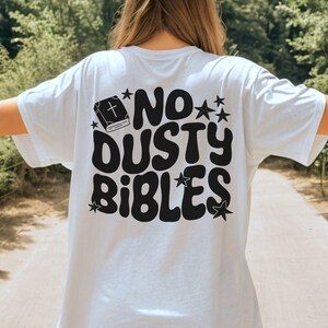 May include: White t-shirt with the phrase "NO DUSTY BIBLES" in black, retro-style lettering. The design includes a book with a cross and star accents. The shirt is a casual, everyday wear item.