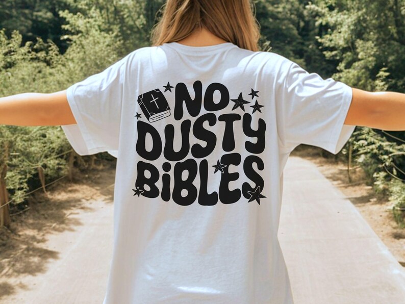 May include: White t-shirt with the phrase "NO DUSTY BIBLES" in black, retro-style lettering. The design includes a book with a cross and star accents. The shirt is a casual, everyday wear item.