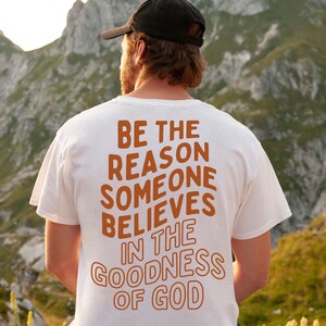 Może przedstawiać: Biały T-shirt z nadrukiem "BE THE REASON SOMEONE BELIEVES IN THE GOODNESS OF GOD" w kolorze pomarańczowym na plecach. Osoba w tym T-shircie stoi przed pasmem g&oacute;rskim.
