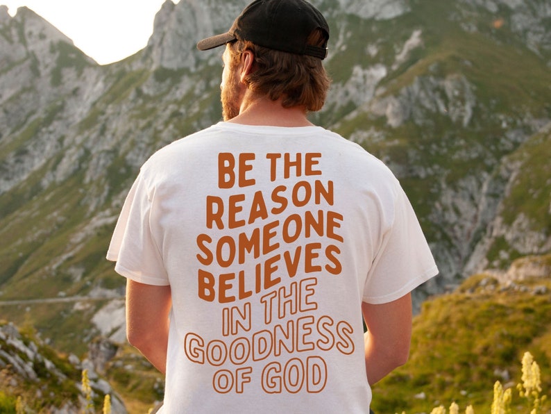 Może przedstawiać: Biały T-shirt z nadrukiem "BE THE REASON SOMEONE BELIEVES IN THE GOODNESS OF GOD" w kolorze pomarańczowym na plecach. Osoba w tym T-shircie stoi przed pasmem g&oacute;rskim.
