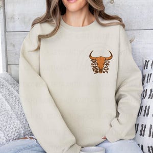May include: Cream-colored sweatshirt featuring a brown bull skull graphic with the text "PSALM 50:10." The design is centered on the chest. The sweatshirt is a neutral color, suitable for various outfits.
