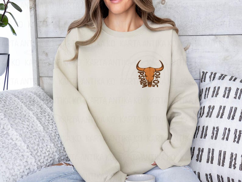 May include: Cream-colored sweatshirt featuring a brown bull skull graphic with the text "PSALM 50:10." The design is centered on the chest. The sweatshirt is a neutral color, suitable for various outfits.