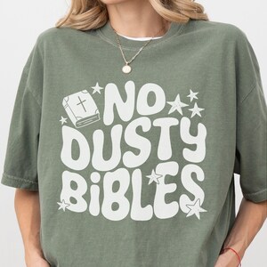 May include: Olive green t-shirt with the text "NO DUSTY BIBLES" in white, along with a graphic of a book and stars. The shirt has short sleeves and a relaxed fit.