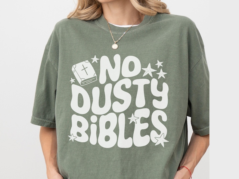 May include: Olive green t-shirt with the text "NO DUSTY BIBLES" in white, along with a graphic of a book and stars. The shirt has short sleeves and a relaxed fit.
