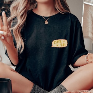 May include: Black t-shirt with a small, yellow speech bubble graphic that reads "ESTER 4:14" in white text. The shirt is worn by someone sitting cross-legged, with a peace sign gesture.