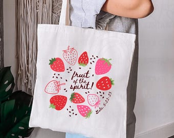 May include: White tote bag featuring a strawberry design with red, pink, and white strawberries arranged in a circle. The text "Fruit of the Spirit!" and "Gala 5:22-23" are also printed on the bag. The bag has a simple, clean design.
